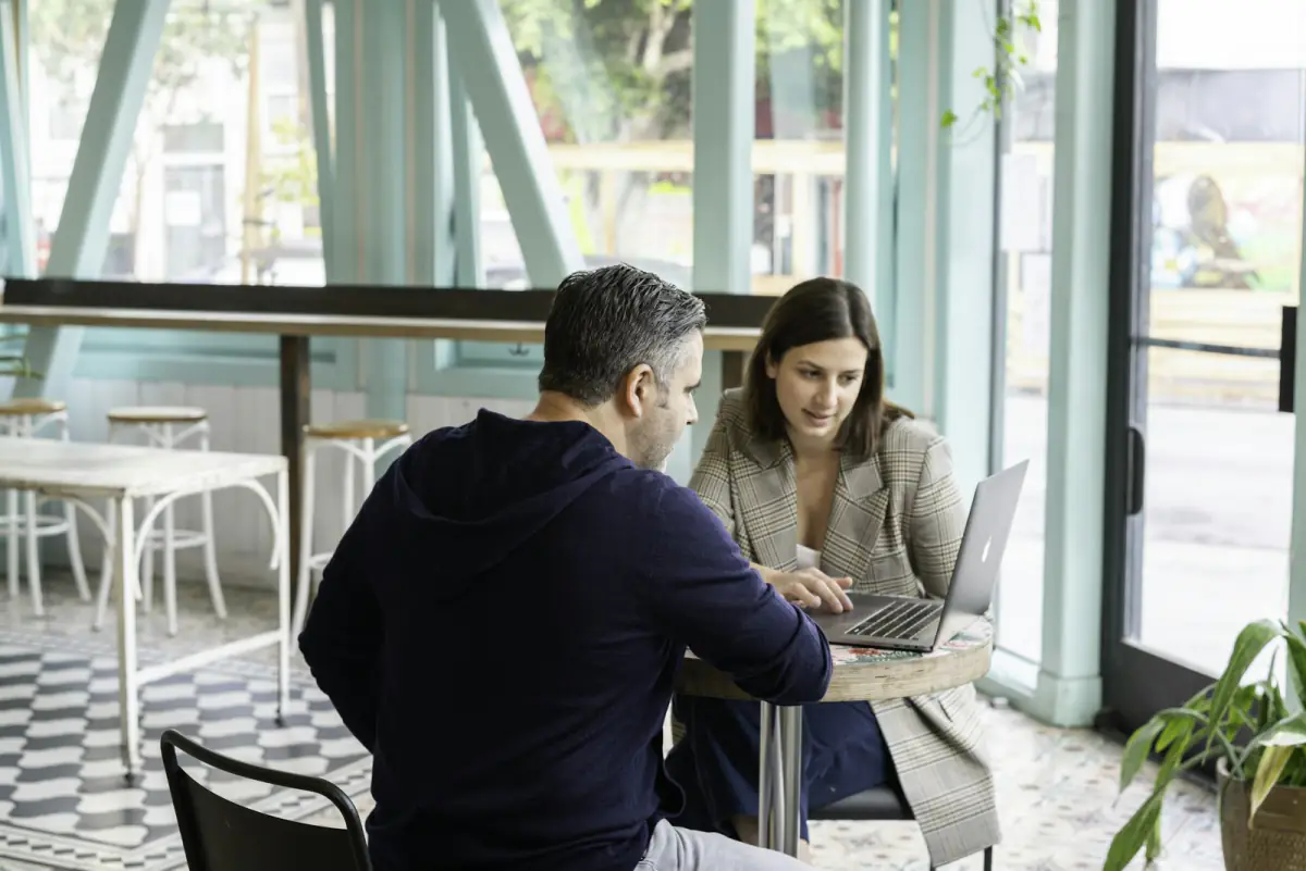 Zwei Menschen besprechen sich an einem Cafétisch über einem Laptop.