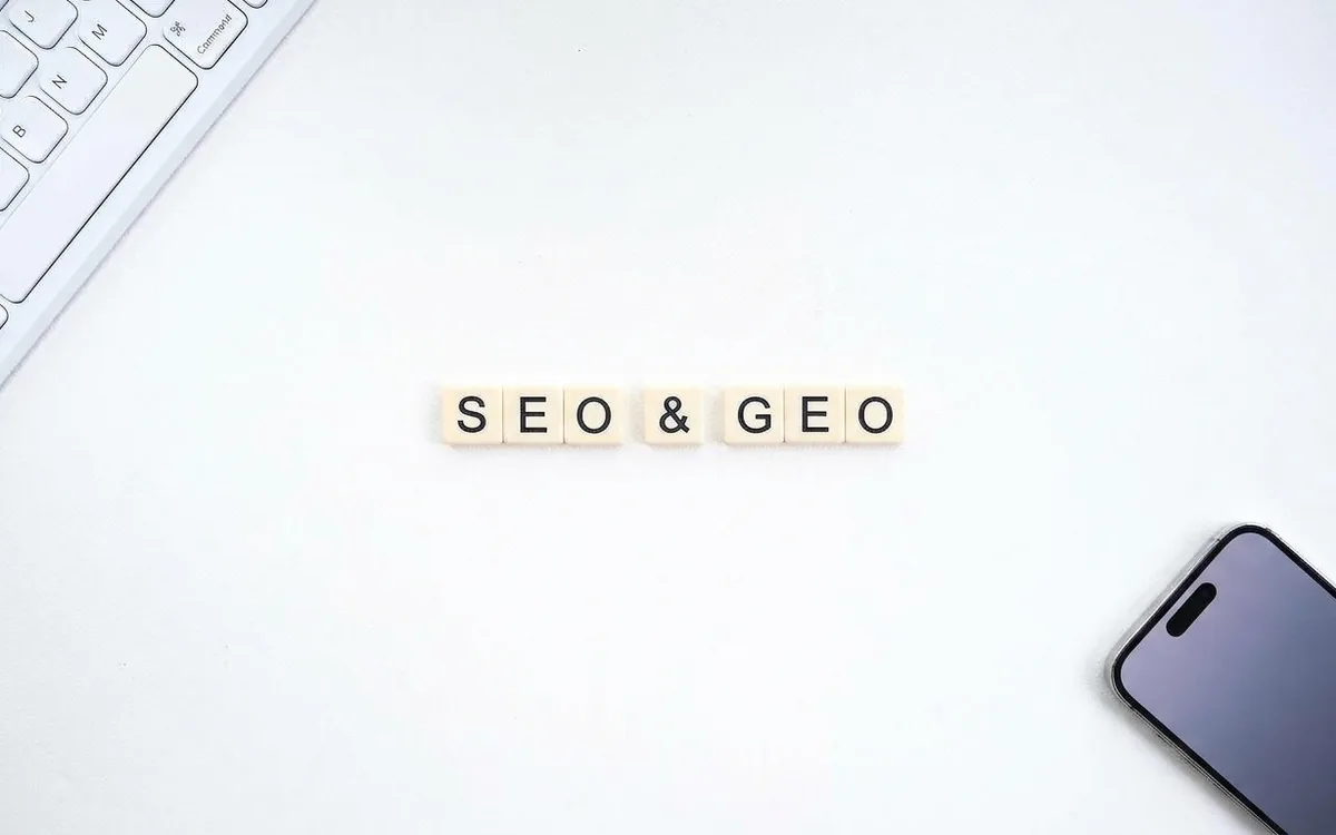 Buchstabensteine auf einem hellen Schreibtisch ergeben das Wort SEO & GEO, daneben eine Tastatur und ein Smartphone.